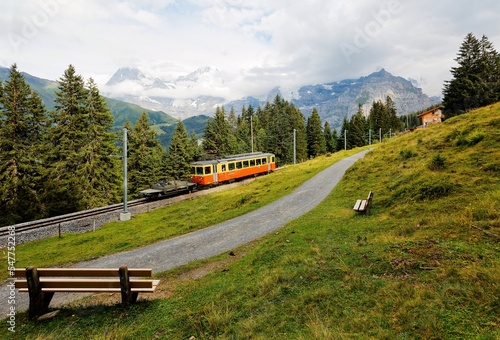 Scenery of a train traveling on a green hillside with wooden benches by a hiking trail and majestic Eiger, Monch and Jungfrau mountains in background, in Murren, Bernese Oberland, Switzerland