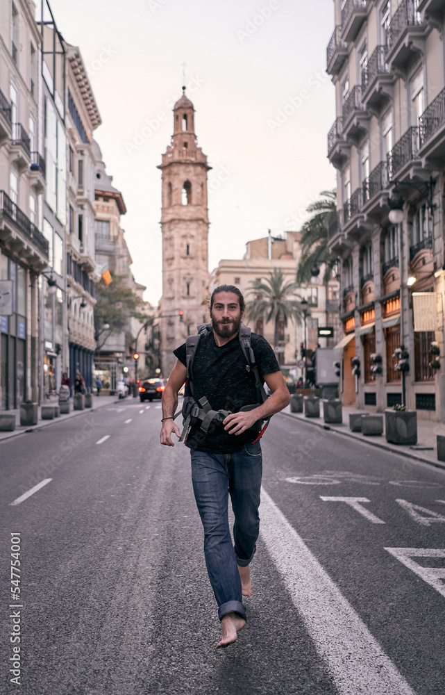 young caucasian man with big backpack on his back running barefoot towards camera through a city street, valencia, spain