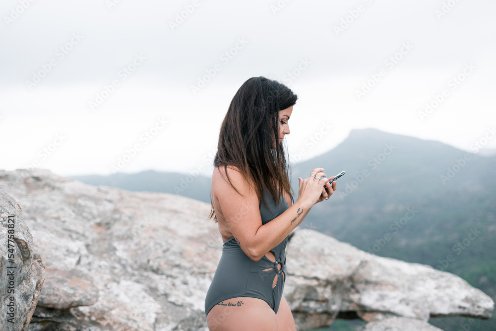 Caucasian mature black haired brunette woman in a gray swimsuit smiling touching the screen of her smartphone