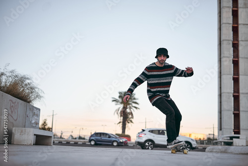caucasian boy in striped sweater with nose piercing and cute beanie having fun on a skateboard in the city at sunset on a clear day