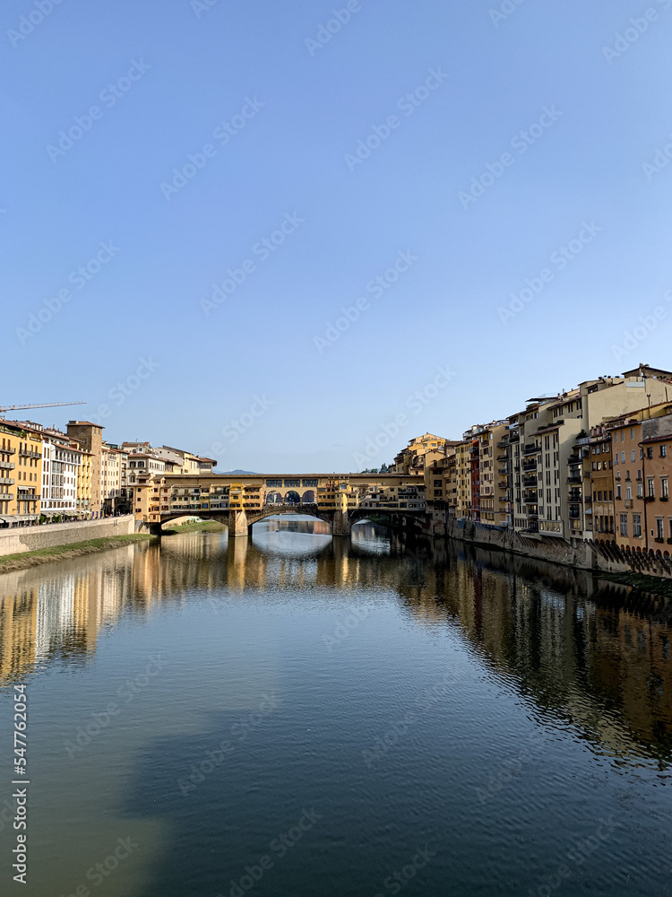Firenze, Tuscany, Italy. Ponte Vecchio Bridge during beautiful sunny day with reflection in Arno River, Florence, Italy. Picturesque medieval arched river bridge with Roman origins, lined
