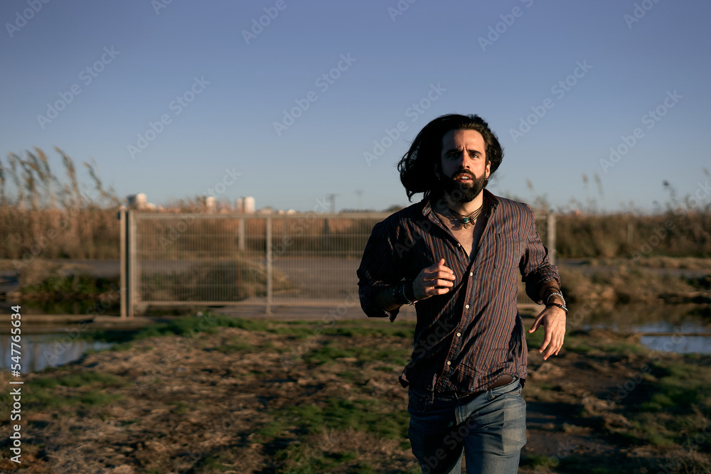 caucasian young man with long hair running towards camera near the metal fence on the outskirts of the city