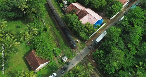 An old train moves through a village in a beautiful green jungle on a bright sunny day. Cars are waiting at the railway crossing for the passage of the train. Top view from drone flying over a train