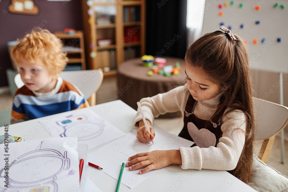 Little learner of nursery school drawing picture with crayons on paper while sitting by desk against gingerhead schoolboy in classroom Little learner of nursery school drawing picture with crayons on paper while sitting by desk against gingerhead schoolboy in classroom