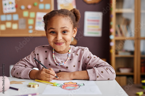 Cute successful learner of primary school looking at camera with smile while sitting by table and drawing with crayons at lesson