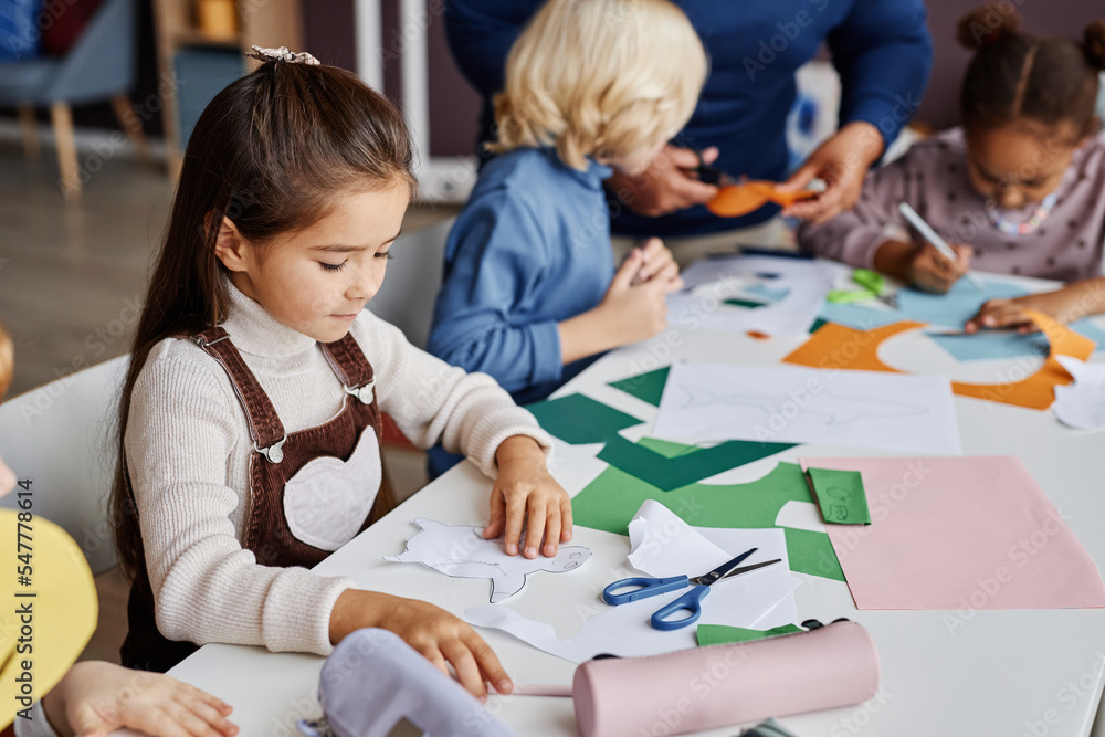 Cute diligent learner of nursery school creating paper toy while sitting by desk among her classmates and teacher at lesson in kindergarten Cute diligent learner of nursery school creating paper toy while sitting by desk among her classmates and teacher at lesson in kindergarten
