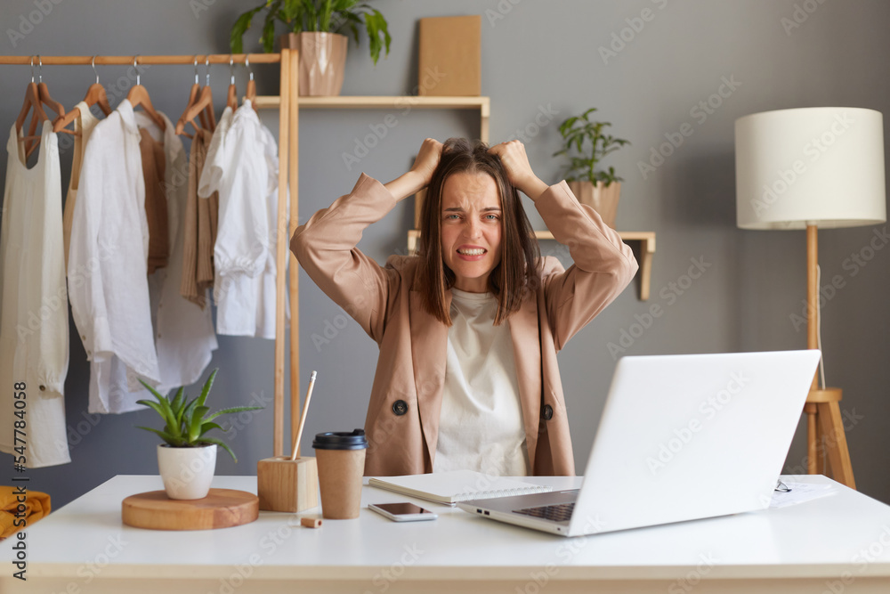 Image of aggressive angry Caucasian brown-haired woman stylist wearing beige jacket sitting at her workplace in front of notebook, screaming, keeping hands on head. Image of aggressive angry Caucasian brown-haired woman stylist wearing beige jacket sitting at her workplace in front of notebook, screaming, keeping hands on head.