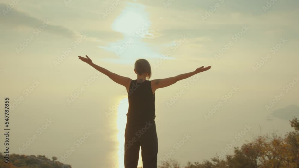Woman raises up her hands while standing in mountains by ocean at sunset. Concept of success and healthy lifestyle Woman raises up her hands while standing in mountains by ocean at sunset. Concept of success and healthy lifestyle