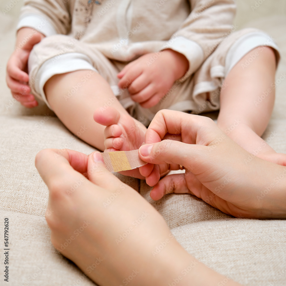 Mother woman sticks a medical band-aid on the toddler baby leg. Mom s hand with sticky wound protection tape and child s foot. Kid aged one year and three months