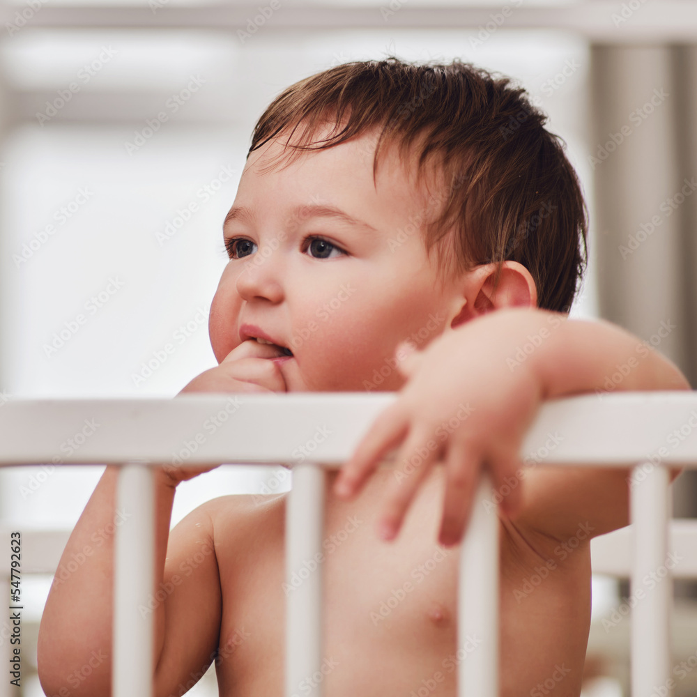 Toddler baby stands thinking in the crib, closeup portrait. The child bites his nails on