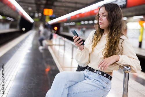 Portrait of a focused girl sitting on a subway platform bench, texing with friends on a mobile phone photo