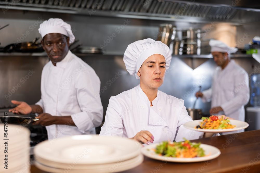 Portrait of focused young female chef cook handing plate with ready meal at restaurant kitchen