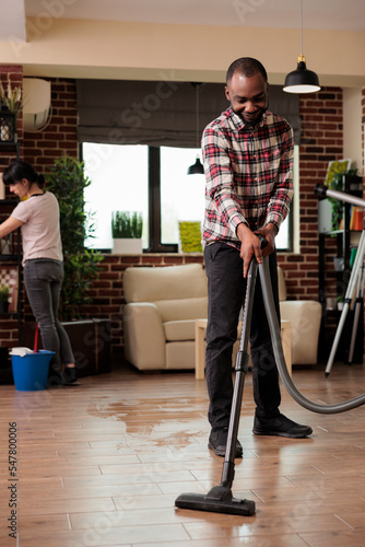 Smiling African American man vacuuming the floor while woman dusts the shelves. Multiracial couple cleaning the house on a saturday morning, doing the weekly cleaning routine. Household chores.