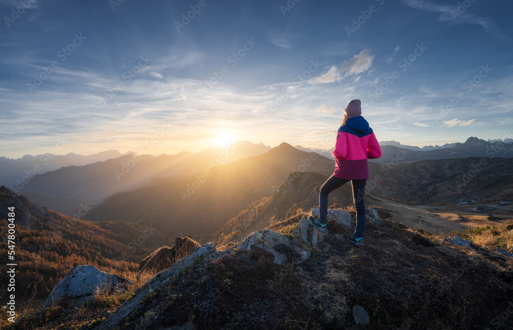 Fototapeta premium Young woman on mountain peak and beautiful mountains in haze at colorful sunset in autumn. Dolomites, Italy. Sporty girl, mountain ridges in fog, orange grass, trees, golden sun in fall. Hiking