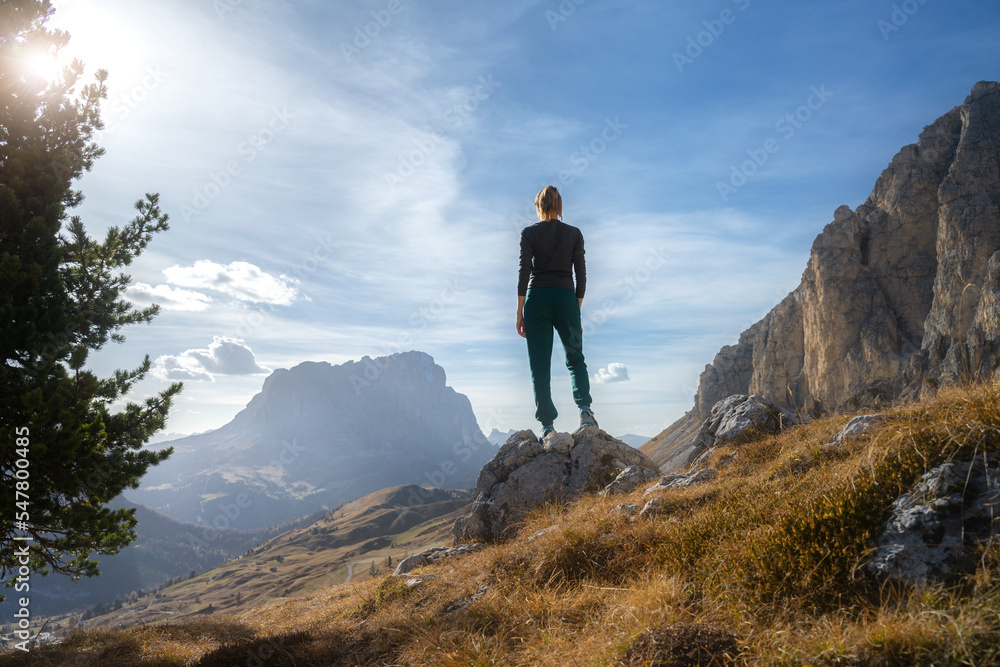 Woman on the stone on mountain trail at sunset in autumn in Dolomites, Italy. Girl on the rock, high mountains, blue sky in fall. Colorful landscape with cliffs, grass. Trekking and hiking. Lifestyle
