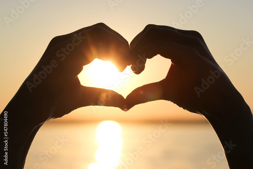 Symbol of love, heart made with fingers of hands on the background of yellow sky at sunset on the background of calm water of the sea, ocean, river. Goodbye summer concept. The end of summer vacation.