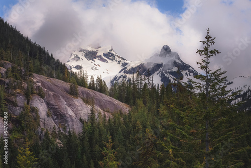 A scenic view of Anif Peak framed by lush pine forests and rugged slopes in British Columbia’s Coast Mountains, under a dramatic, cloud-filled sky. photo