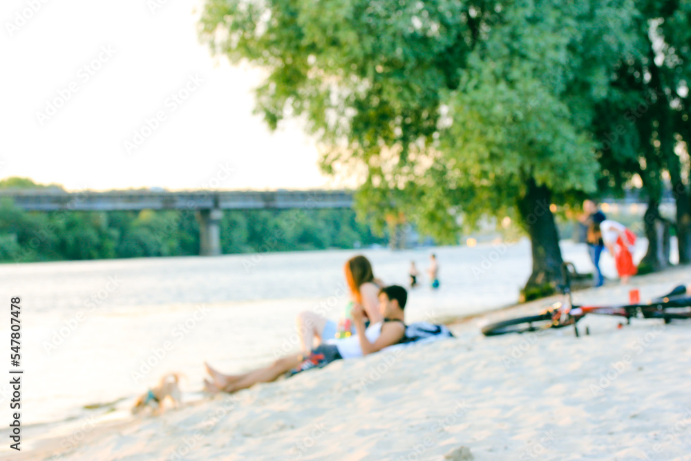 People relaxing on sandy beach, the bank of a river, lake, sea in the summer. Blurred background with people on the street, guy and girl relaxing in nature at the pond on natural green area, park zone