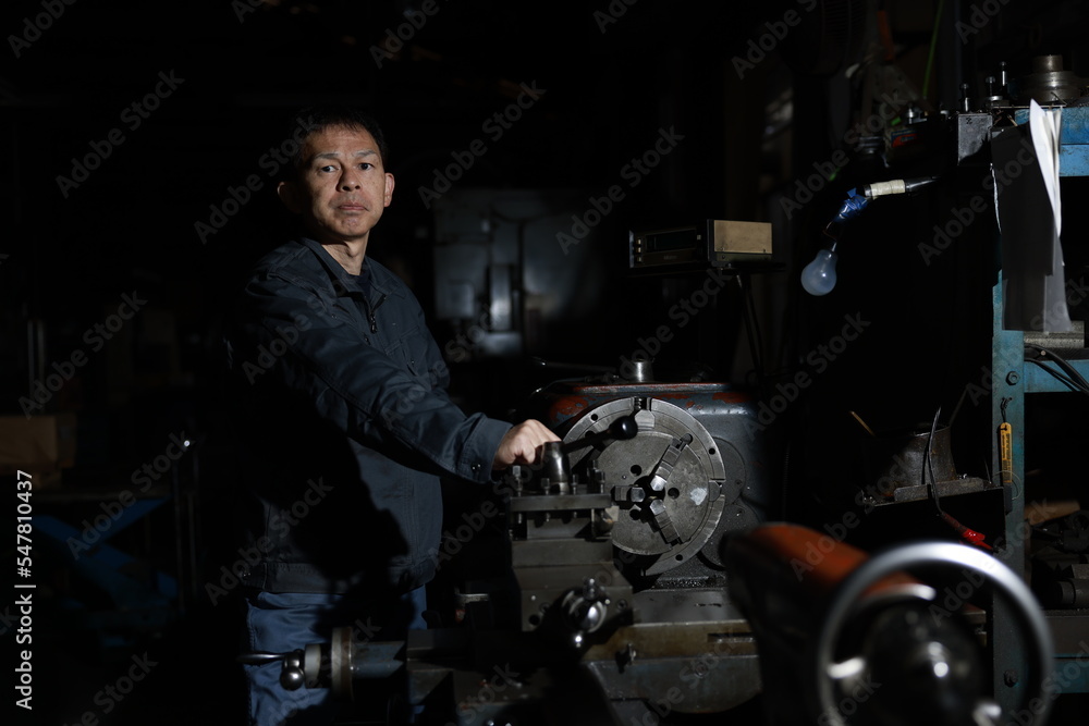 A craftsman poses in gray work clothes in front of a lathe at a local factory. Conceptual images of the essence of manufacturing, technical succession, and the challenge of high-precision machining. A craftsman poses in gray work clothes in front of a lathe at a local factory. Conceptual images of the essence of manufacturing, technical succession, and the challenge of high-precision machining.