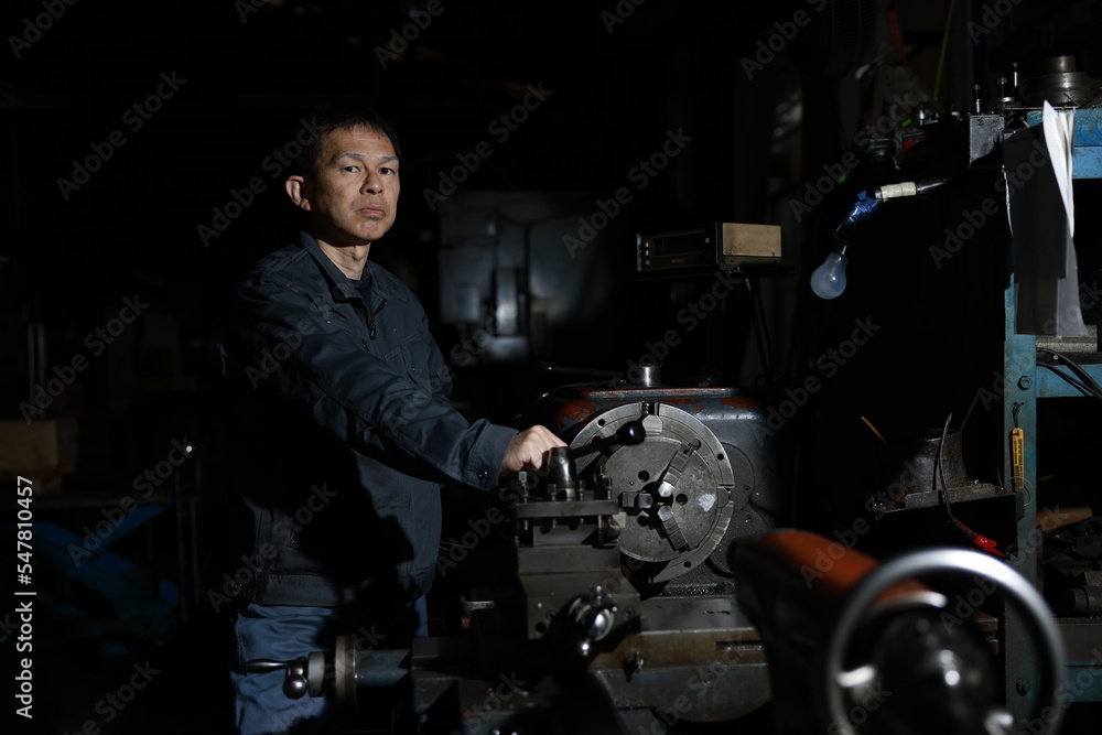 A craftsman poses in gray work clothes in front of a lathe at a local factory. Conceptual images of the essence of manufacturing, technical succession, and the challenge of high-precision machining. A craftsman poses in gray work clothes in front of a lathe at a local factory. Conceptual images of the essence of manufacturing, technical succession, and the challenge of high-precision machining.