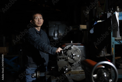 A craftsman poses in gray work clothes in front of a lathe at a local factory. Conceptual images of the essence of manufacturing, technical succession, and the challenge of high-precision machining.