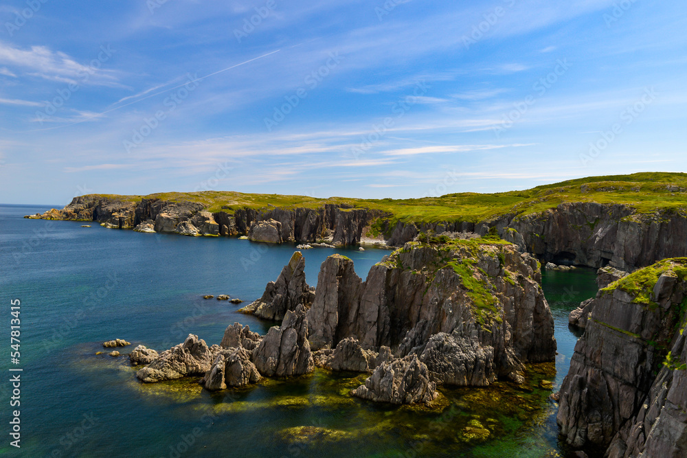 A rugged ocean coastline with high rocky sea stacks, cliffs, vibrant green grass, blue sky, and clouds. The smooth water is a blue color. There's a hiking trail along the edge of the cliff and coast. 