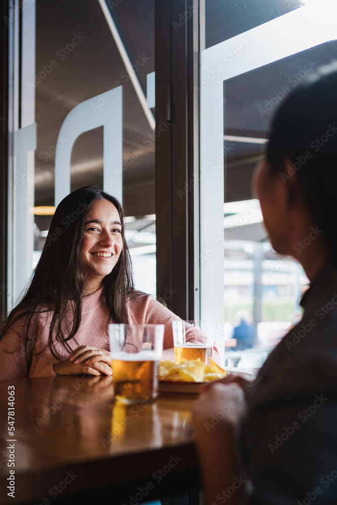 Two smiling girls talking with each other in a bar. They are having some beers and snack Two smiling girls talking with each other in a bar. They are having some beers and snack