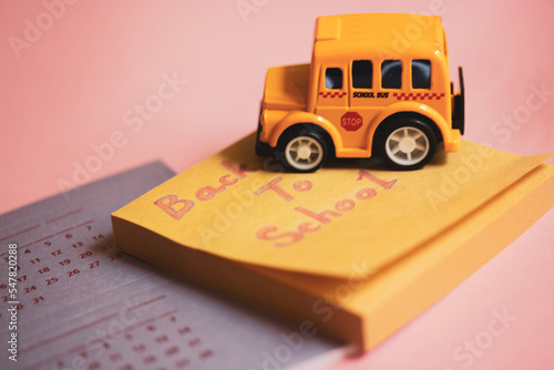 Toy yellow American school bus on table, calendar with dates, colorful wooden pencils lying on pink table. Stationery for studying, drawing, planning. Back to school text on notes. Supplies for study.