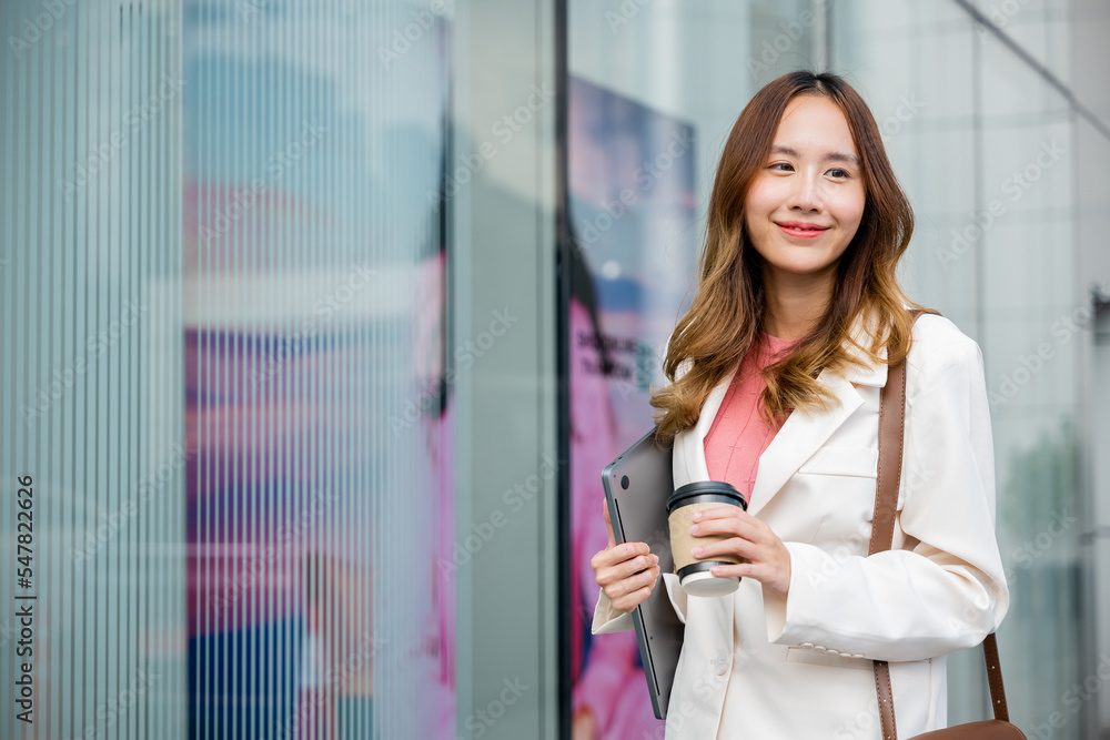 Young business woman wearing white suit jacket and smile while go to work outdoor mirror building background. Confident Businesswoman with cup of coffee holding Laptop walking outside office building Young business woman wearing white suit jacket and smile while go to work outdoor mirror building background. Confident Businesswoman with cup of coffee holding Laptop walking outside office building