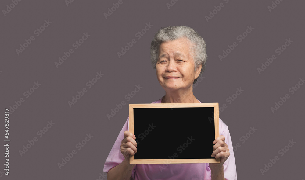An elderly Asian woman holding a mini blackboard and looking at the camera with a smile while standing on a gray background An elderly Asian woman holding a mini blackboard and looking at the camera with a smile while standing on a gray background