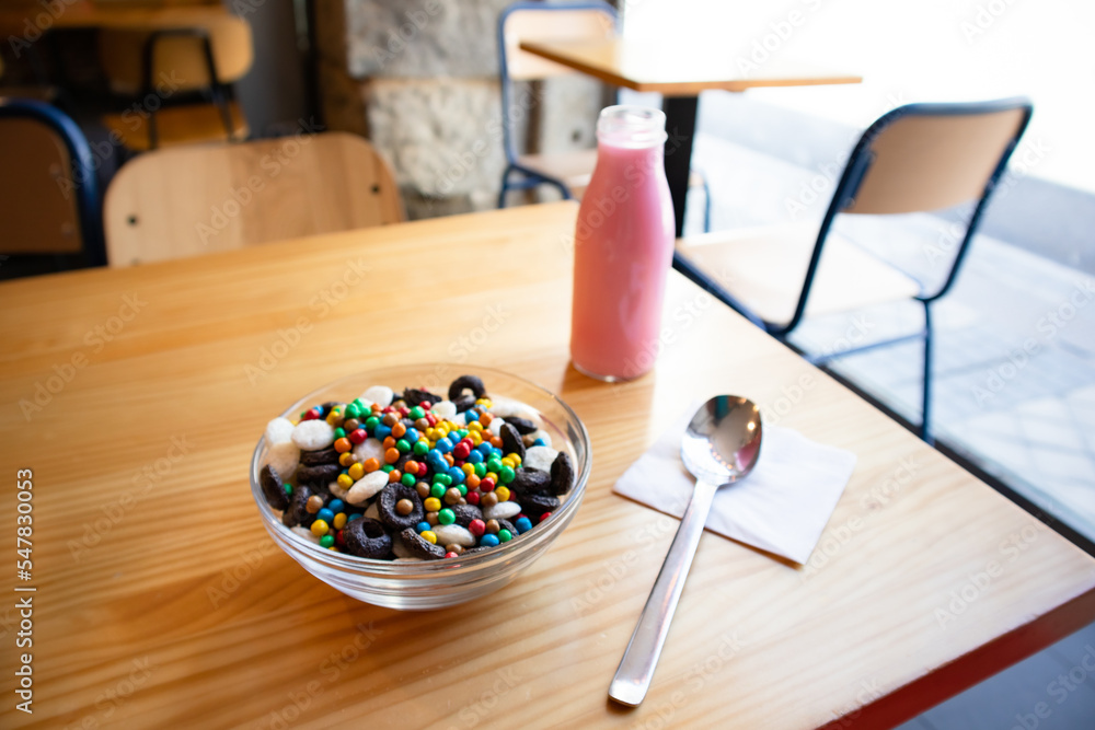 Dry breakfast in glass round plate bowl on a wooden table in cafe. Delicious cornflakes and pink milk in a bottle. Useful healthy breakfast. Food for kids. Different type cereals, multicolored balls. Dry breakfast in glass round plate bowl on a wooden table in cafe. Delicious cornflakes and pink milk in a bottle. Useful healthy breakfast. Food for kids. Different type cereals, multicolored balls.