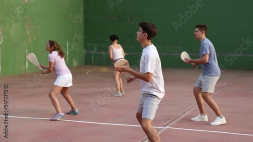 Group of people playing pelota at open-air fronton in summer, swinging traditional wooden bat to return ball. Sportsman ready to hit volley photo