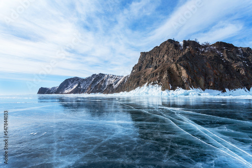 Frozen Baikal Lake on cold February day. View from clear blue ice on rocky coast of Cape Hoboy - a natural landmark of Olkhon Island. Beautiful winter landscape. Ice travel and outdoor recreation