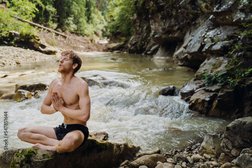 White man wearing swimming trunks meditating while resting by river