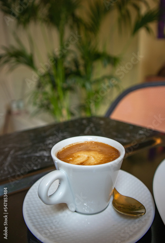 Porcelain white coffee cup with cappuccino, mochaccino on a saucer with a teaspoon on a black marble table to view. Hot tasty drink for a breakfast, branch, lunch. Aromatic beverage on a kitchen.