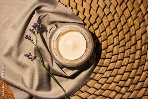 Still life with moisturizer face or body cream in a round jar with a purple flowers top view. Women's skin care. Natural eco cosmetics on rattan background flatly. Silver silk handkerchief Copy space
