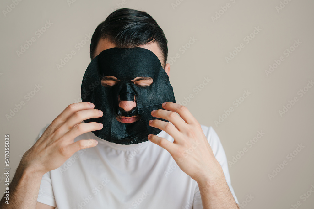Close-up portrait adult man applying a black moisturizing paper mask isolated over gray background. Beauty treatment. Facial skincare. Close-up portrait adult man applying a black moisturizing paper mask isolated over gray background. Beauty treatment. Facial skincare.