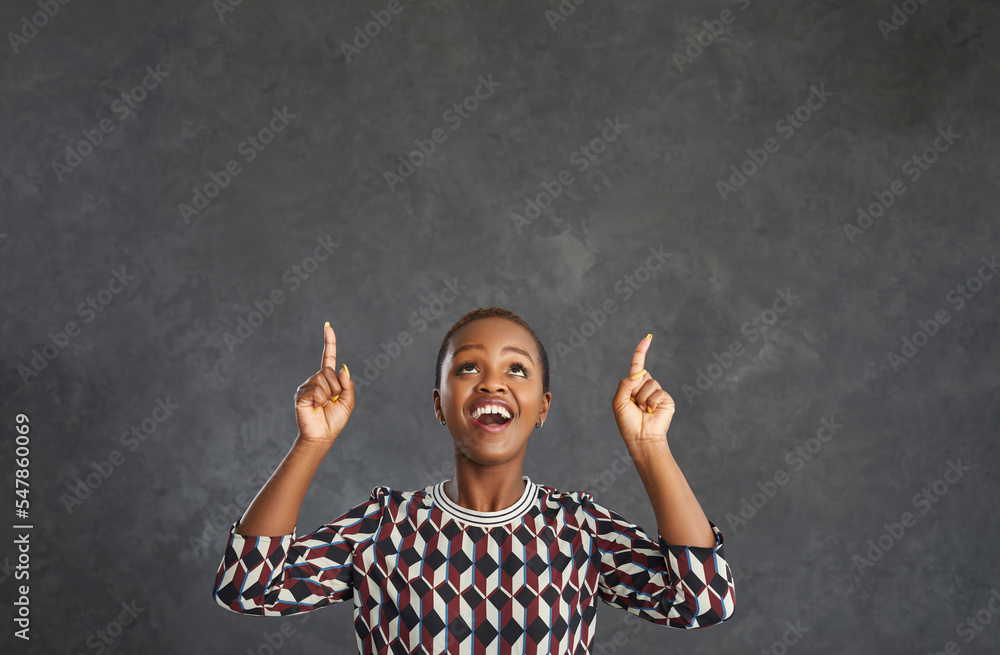 Happy dark skinned woman looking up and pointing her index finger up at a free space for text offers something to choose from. Joyful woman on a background of a gray wall. People and promotion concept Happy dark skinned woman looking up and pointing her index finger up at a free space for text offers something to choose from. Joyful woman on a background of a gray wall. People and promotion concept