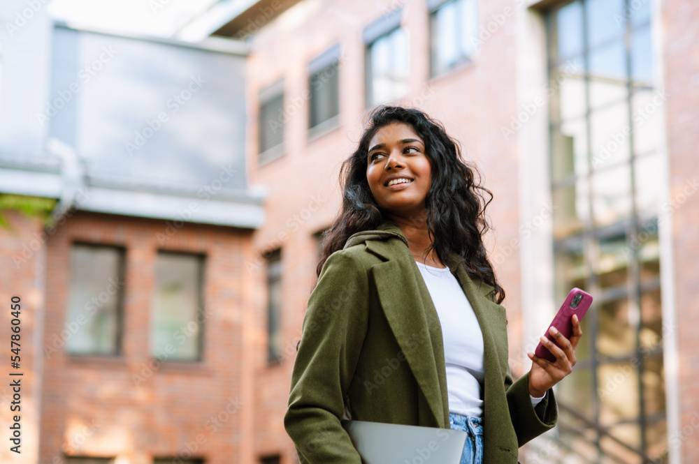 Brunette indian woman using mobile phone while walking on city street