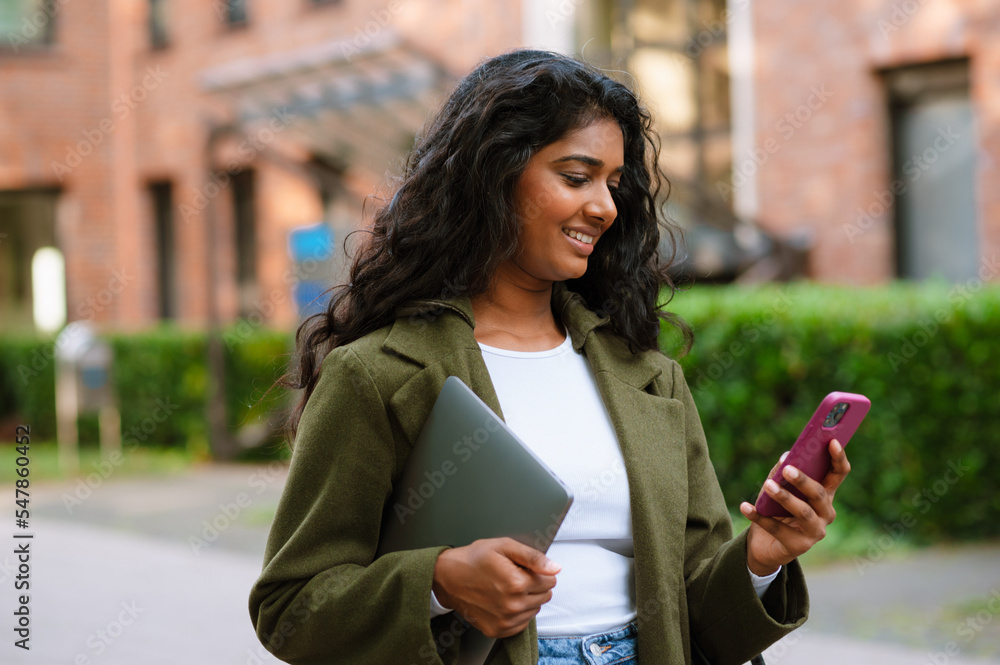 Brunette indian woman using mobile phone while walking on city street