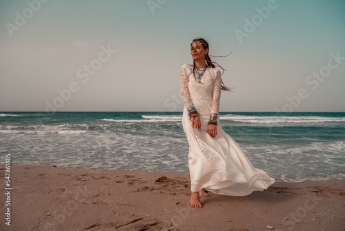 Model in boho style in a white long dress and silver jewelry on the beach. Her hair is braided  and there are many bracelets on her arms.