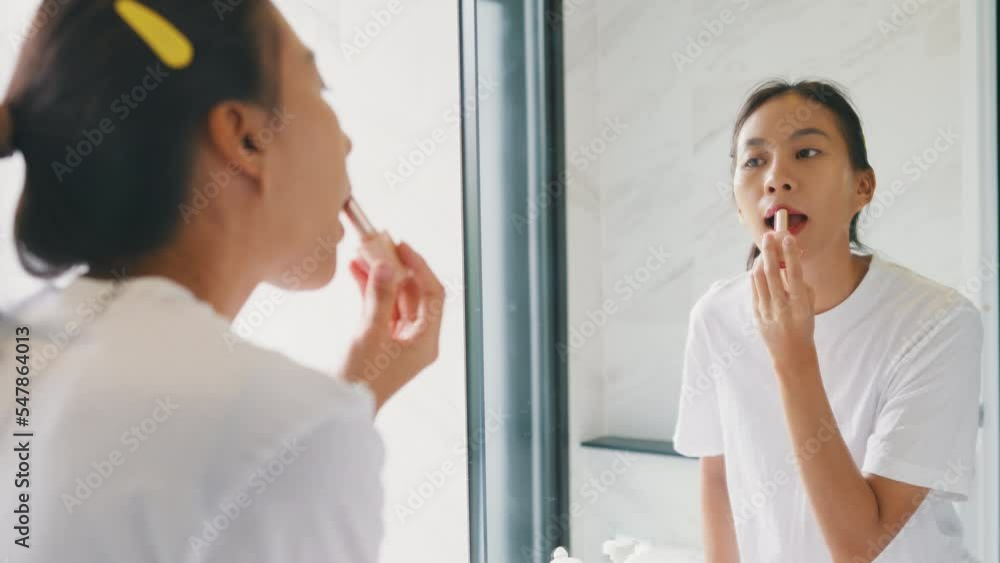 Young Asian happy woman make up looking at her reflection in mirror apply lip balm on her lips in bathroom. Lifestyle and people, Lips Protection, Natural Skin Care Cosmetics, Morning routine concept. Young Asian happy woman make up looking at her reflection in mirror apply lip balm on her lips in bathroom. Lifestyle and people, Lips Protection, Natural Skin Care Cosmetics, Morning routine concept.