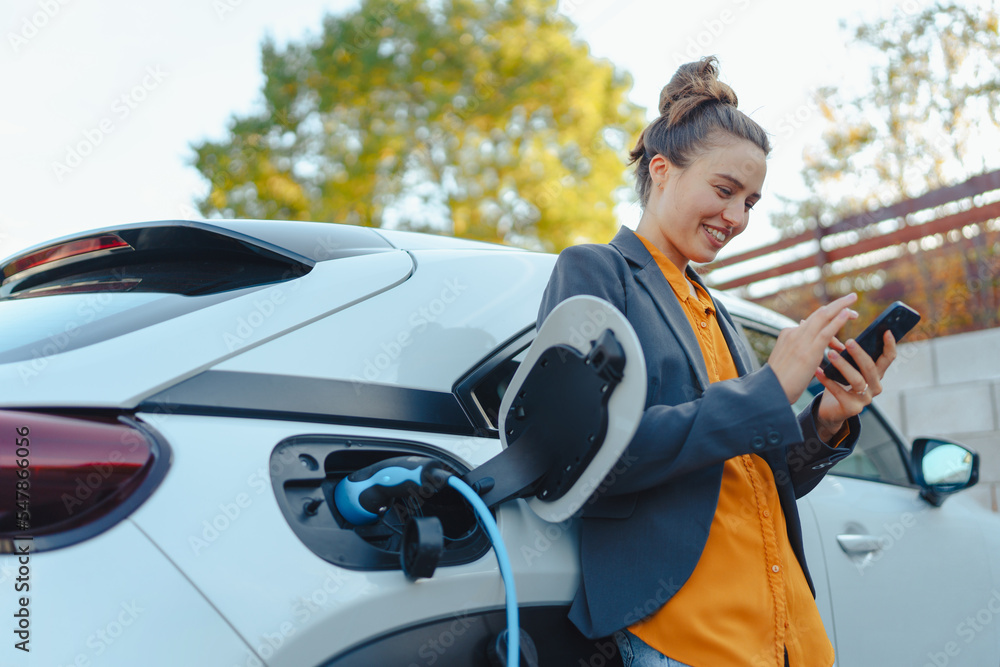 Young woman with smartphone waiting while her electric car charging in home charging station, sustainable and economic transportation concept. - obrazy, fototapety, plakaty 
