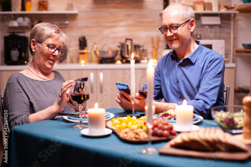 Happy old couple smiling using phone in kitchen having romantic dinner. Sitting at the table in the dining room   browsing  searching  using phone  internet  celebrating their anniversary