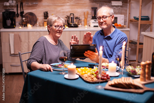Smiling senior couple waving at camera of tablet pc during video call and dinner in kitchen. Couple sitting at the table  browsing  talking  using internet  celebrating their anniversary in dining