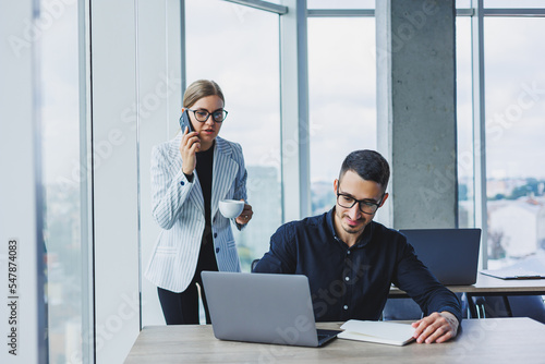 A businessman is working on a laptop while talking while his colleague is listening. Work in a modern bright office