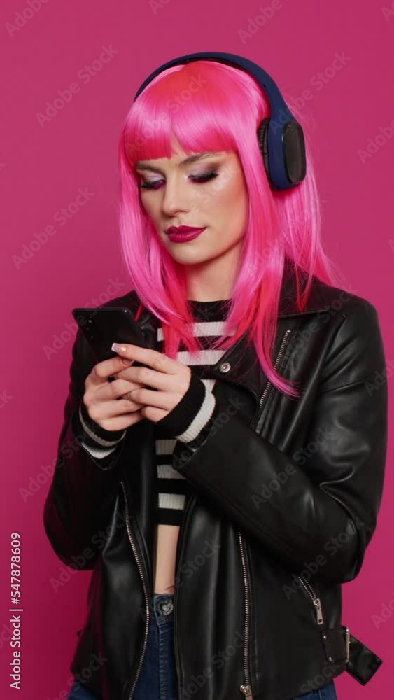 Vertical video: Cheerful confident adult with pink hair texting messages, listening to music on headset. Cool stylish woman having fun with mp3 songs, using social media online app on smartphone. Vertical video: Cheerful confident adult with pink hair texting messages, listening to music on headset. Cool stylish woman having fun with mp3 songs, using social media online app on smartphone.