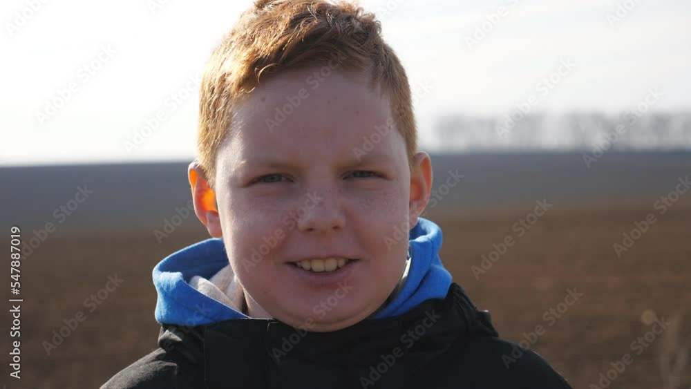 Portrait of little red-haired boy looks into camera with angry facial expression against the blurred background of plowed field. Small annoyed male child with freckles stands on ploughed meadow Portrait of little red-haired boy looks into camera with angry facial expression against the blurred background of plowed field. Small annoyed male child with freckles stands on ploughed meadow