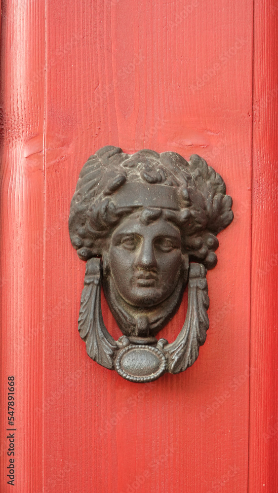 Cast doorknob in the shape of an ancient Roman woman's head on a red door. Antique door knocker