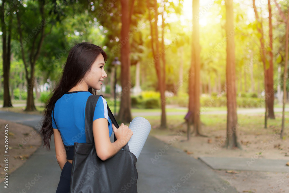 Beautiful woman wearing blue workout clothes holding rolled-up exercise mat in the park evening time. She is happy to go to the exercise. Beautiful woman wearing blue workout clothes holding rolled-up exercise mat in the park evening time. She is happy to go to the exercise.